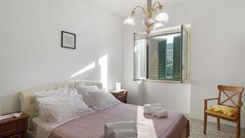 A bedroom with a bed, a nightstand, a chair, a window with shutters, and a chandelier.