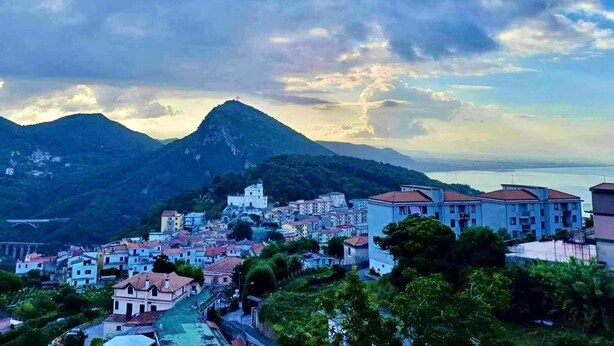 Una località costiera con una montagna imponente sullo sfondo, edifici residenziali e un cielo sereno con nuvole.