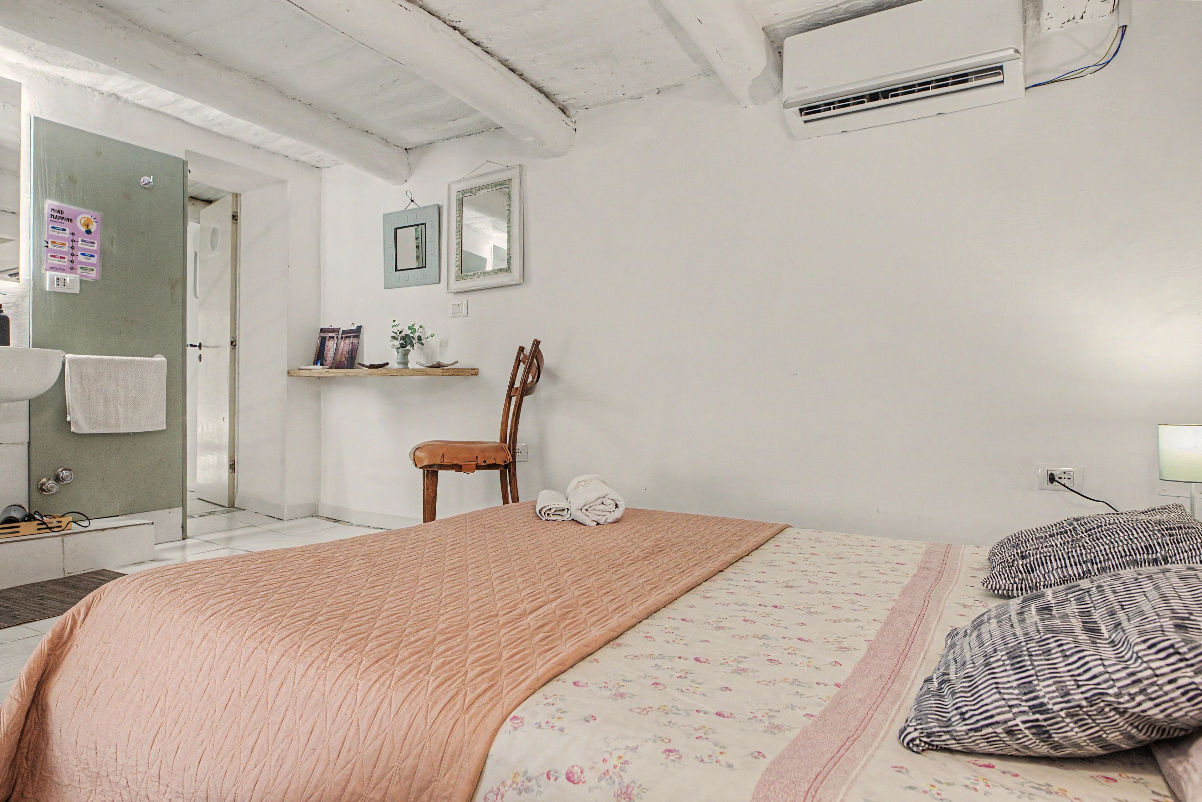 Un dormitorio con cama, una silla, mesita de noche, espejo y unidad de aire acondicionado.