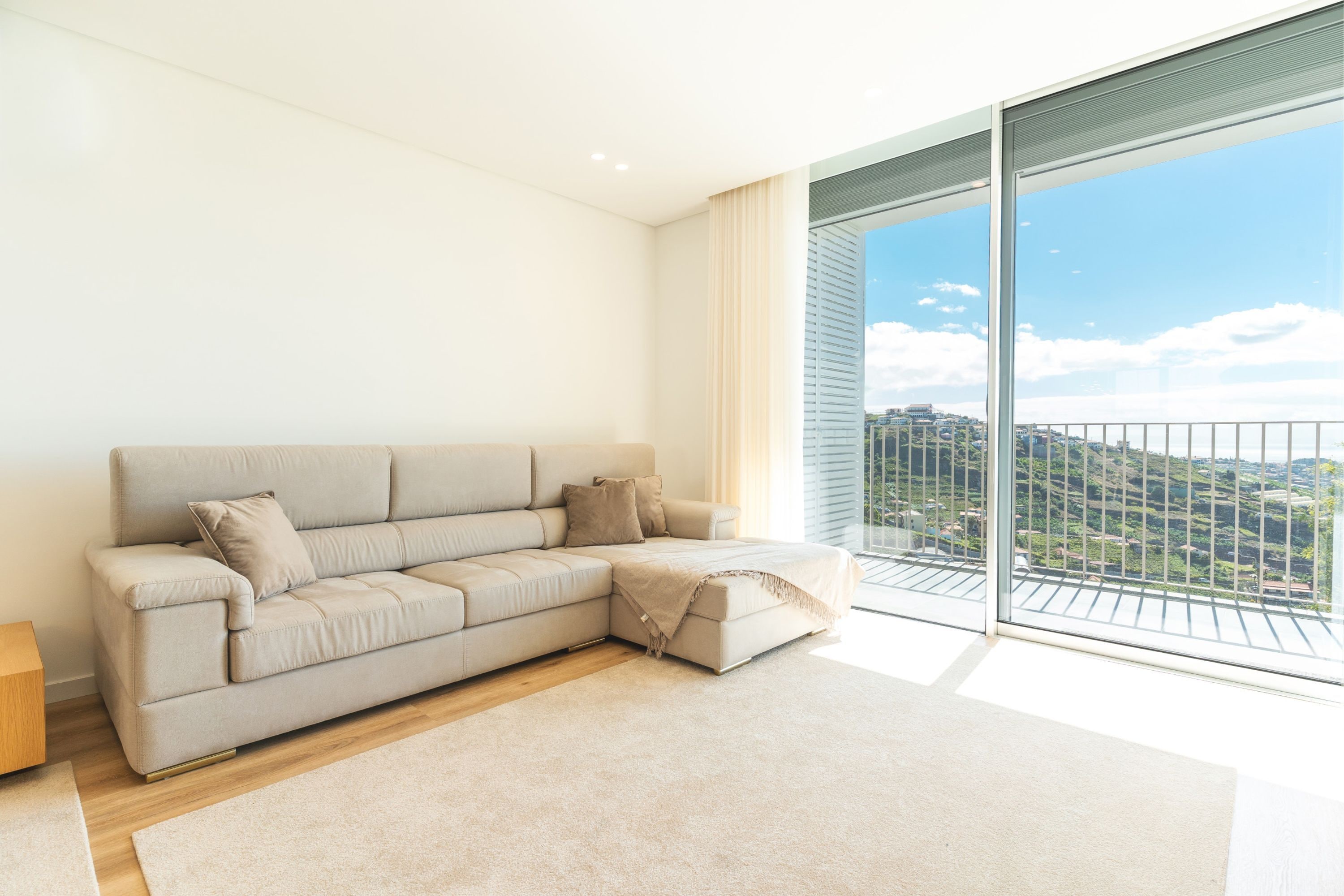 Ein modernes Wohnzimmer mit einem beigen Ecksofa, Holzboden und einem großen Schiebeglastür, die auf einen Balkon mit Blick auf die Stadt führt.