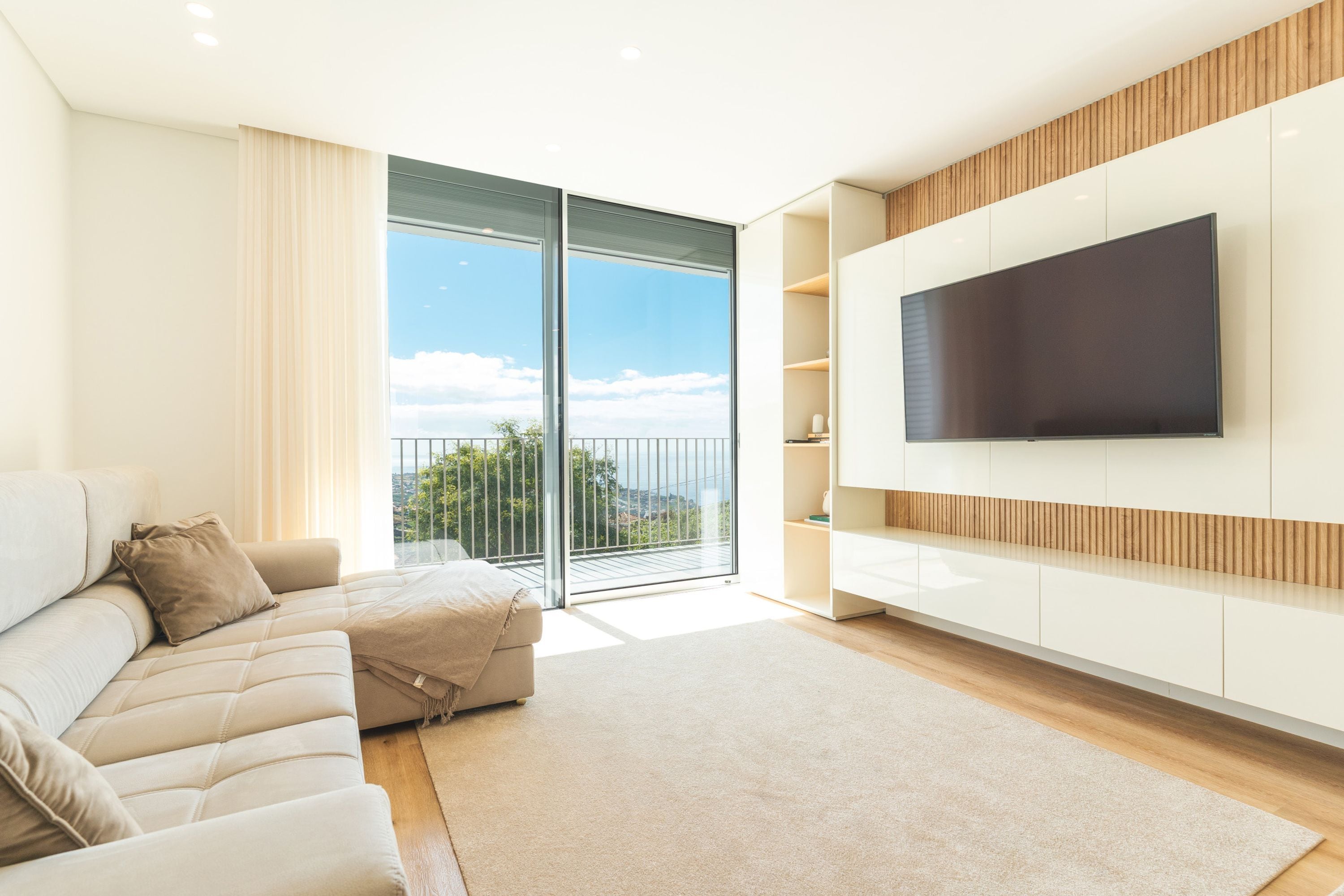 A modern living room with a sofa, a flat-screen TV mounted on the wall, and a sliding glass door leading to a balcony.