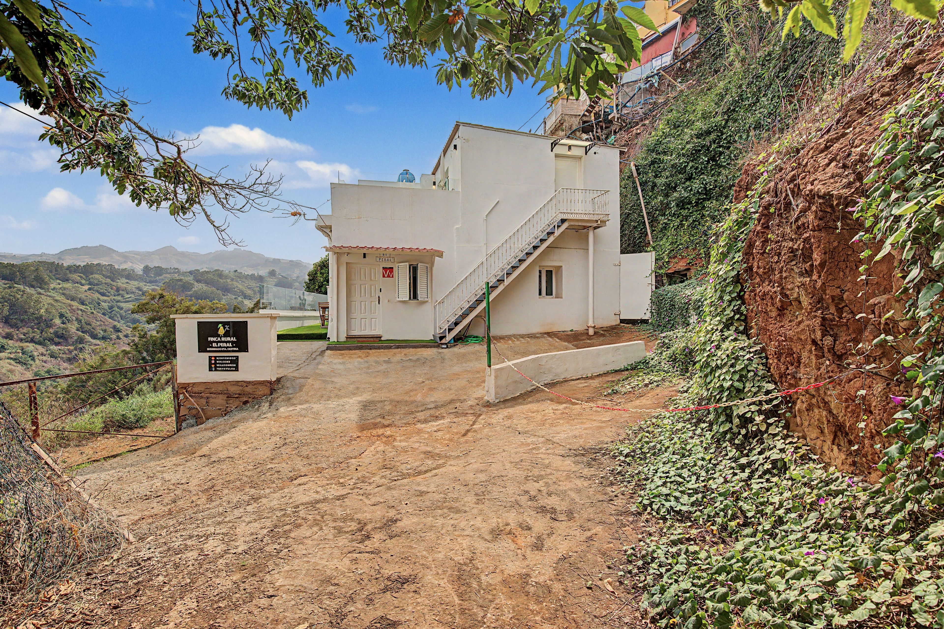 A building with a staircase leading to a door, surrounded by greenery and a dirt path.