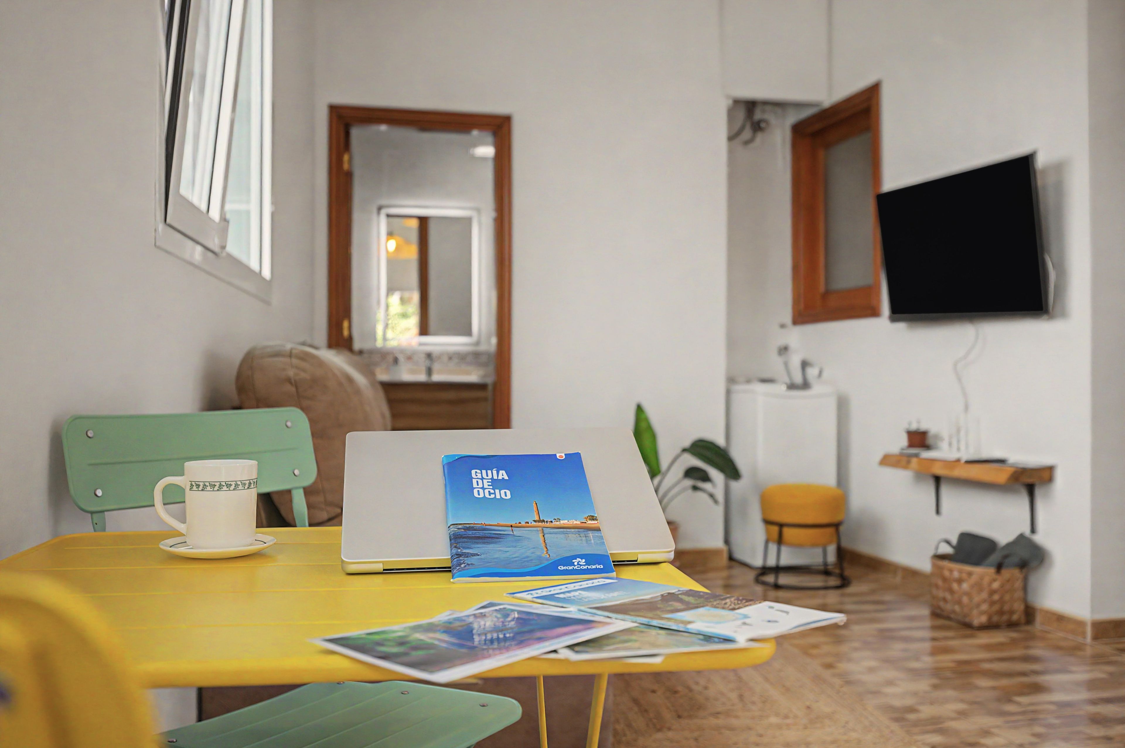 Una habitación con una mesa amarilla, una computadora portátil, una guía de viaje, una taza y un televisor en la pared.