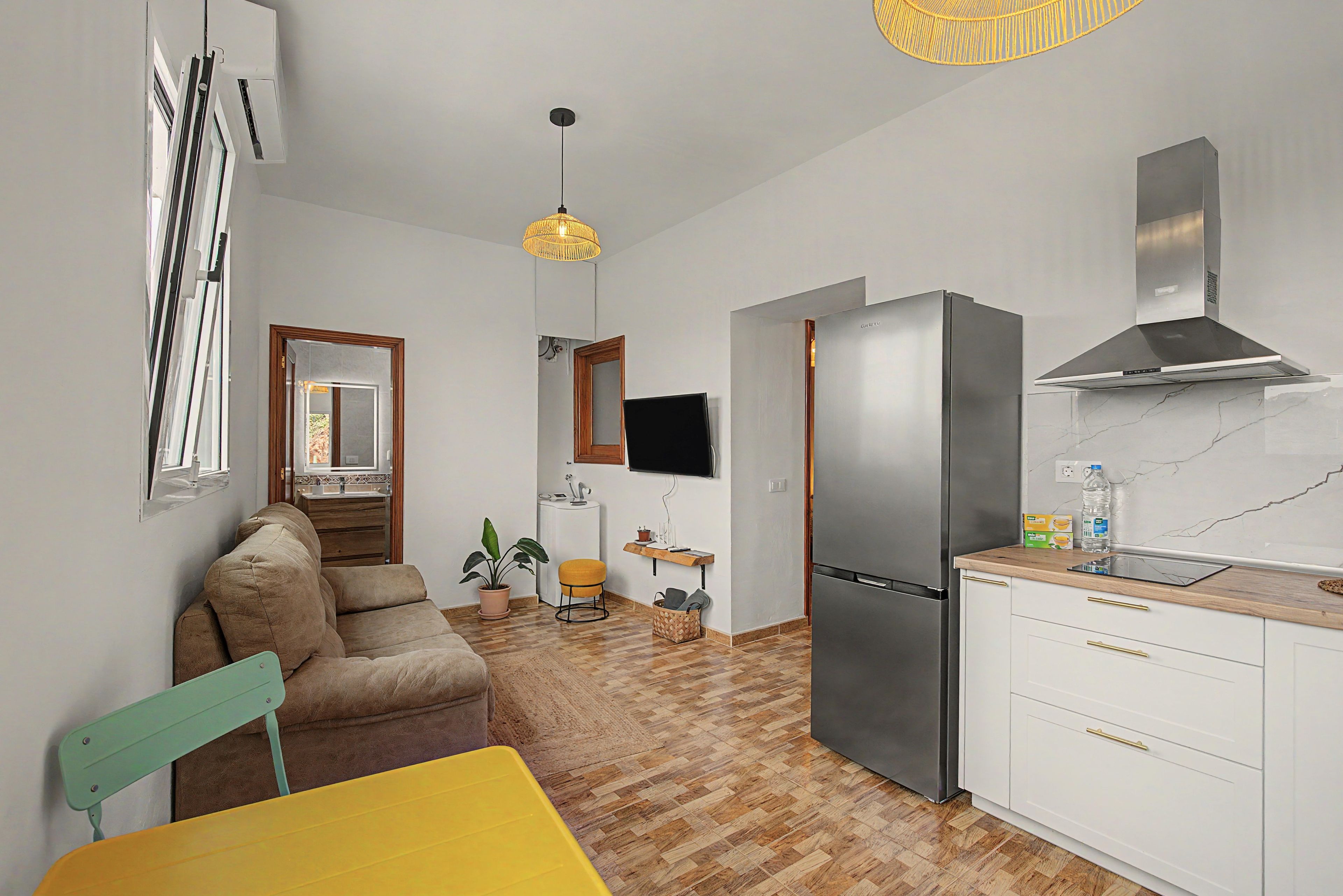 Cocina compacta con un refrigerador moderno, un comedor con una mesa amarilla y una zona de estar con un sofá marrón.