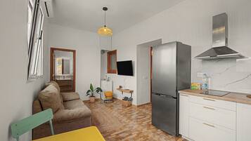 Cocina compacta con un refrigerador moderno, un comedor con una mesa amarilla y una zona de estar con un sofá marrón.
