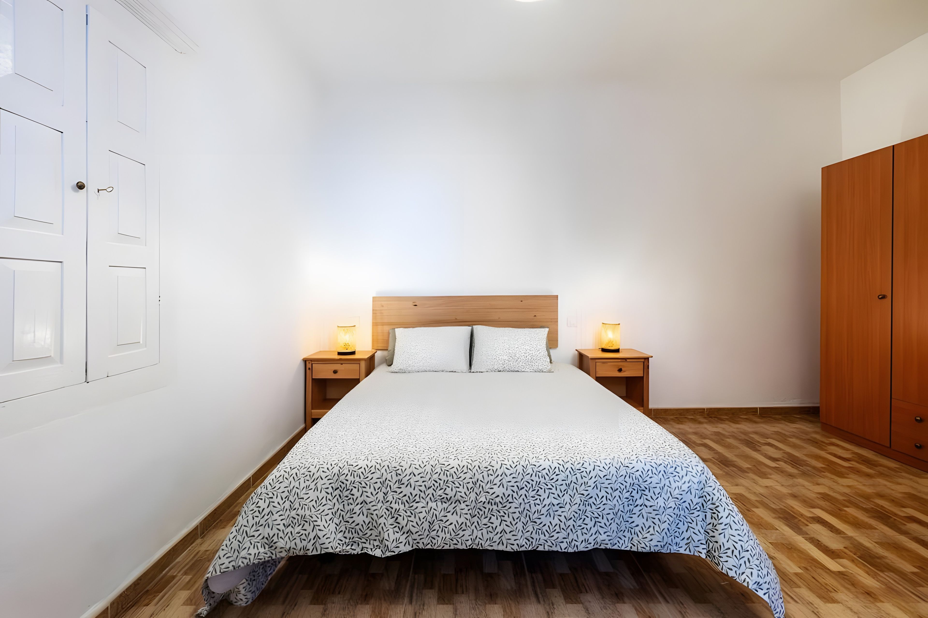 Une chambre à coucher avec un lit, deux tables de chevet avec des lampes, une armoire en bois et un mur blanc avec une porte.