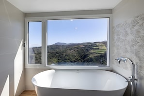 Ein modernes Badezimmer mit einer großen Badewanne und zwei Fenstern mit Aussicht auf Hügel und Grünland.