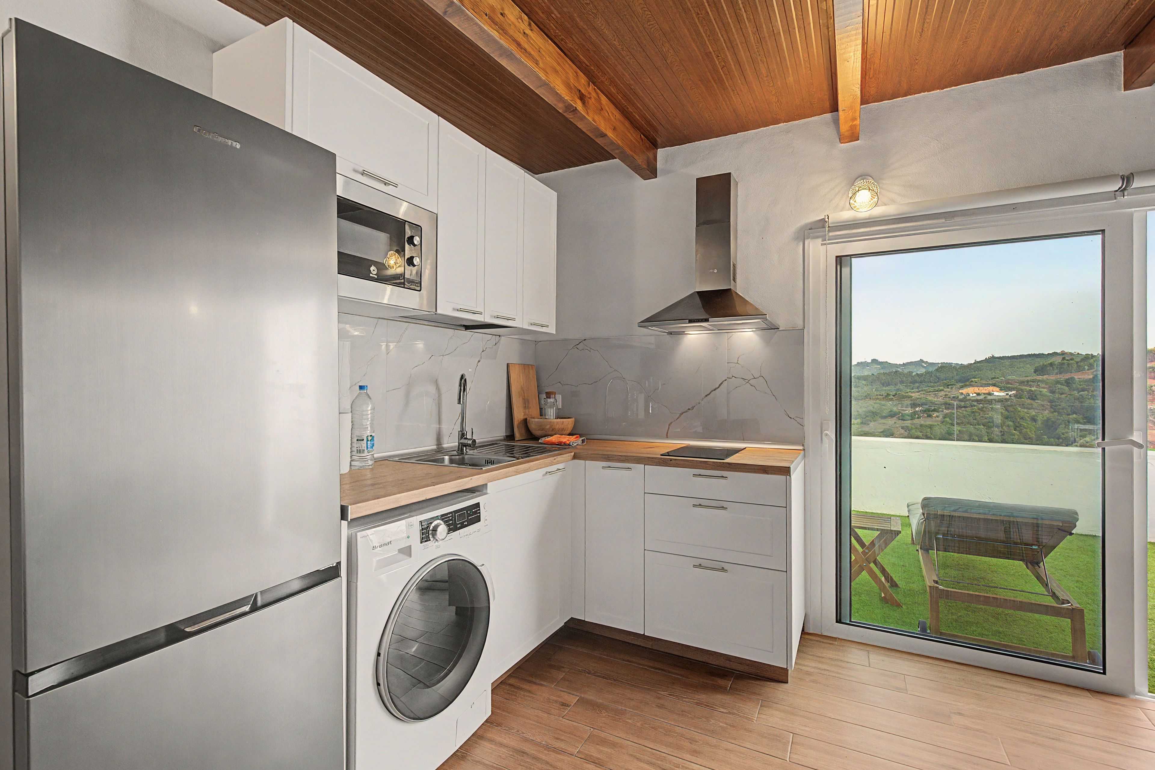 A modern kitchen with a built-in washer, a wooden ceiling, and a view of a green landscape through a glass door.