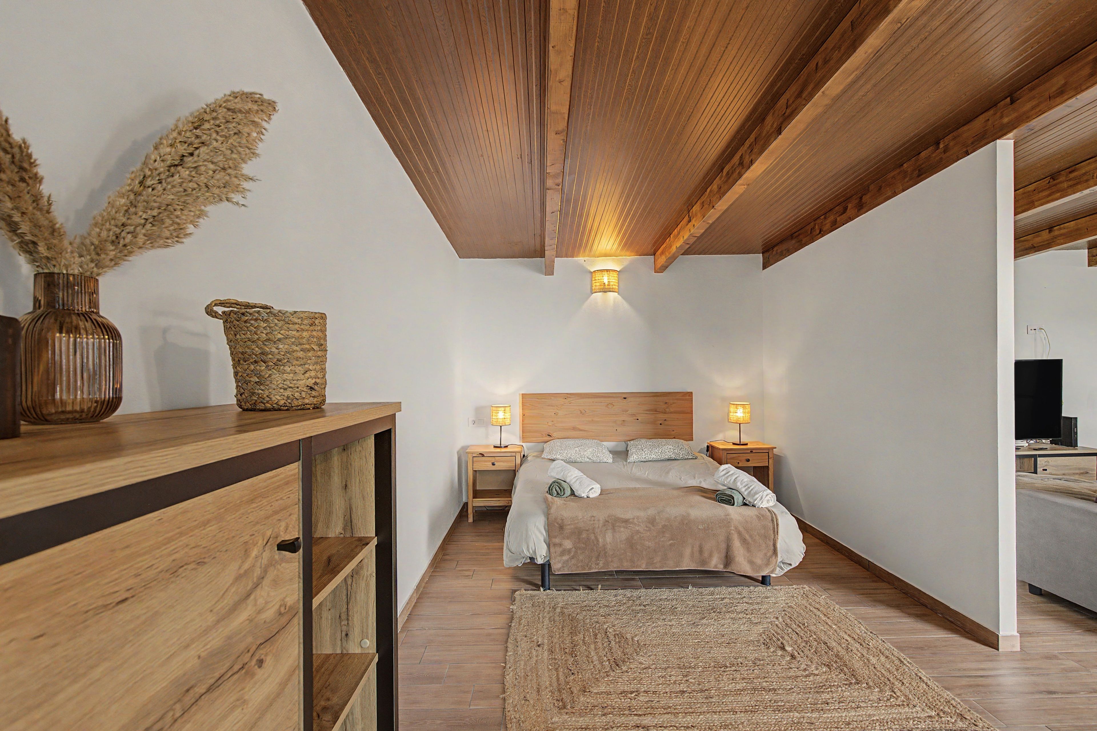 Un dormitorio con techo de madera, una cama con cabecera, mesitas de noche con lámparas, un aparador de madera y una cesta tejida.
