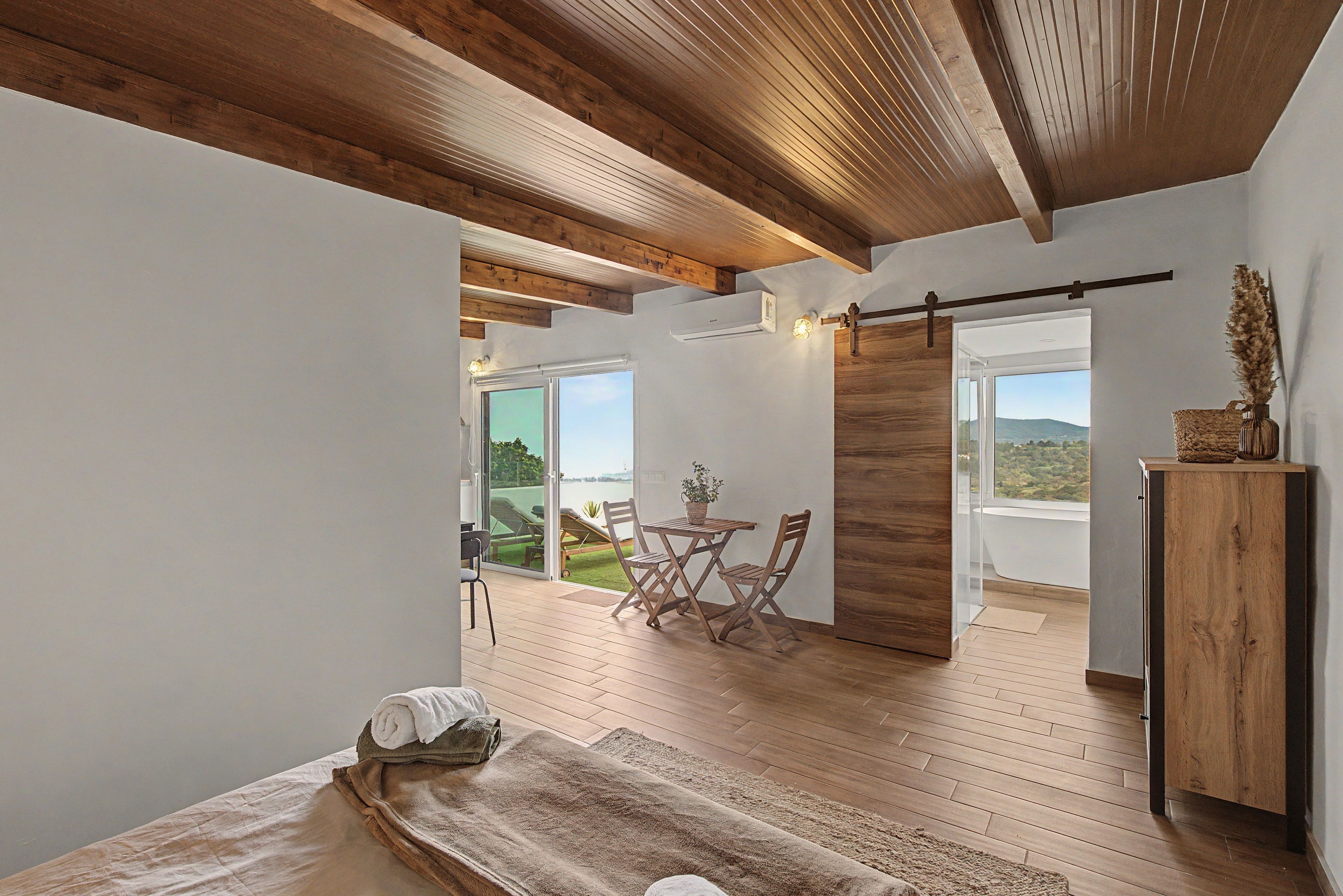 Una habitación con techo de madera, un ventanal grande, una cama con toalla doblada y vistas a un paisaje verde.