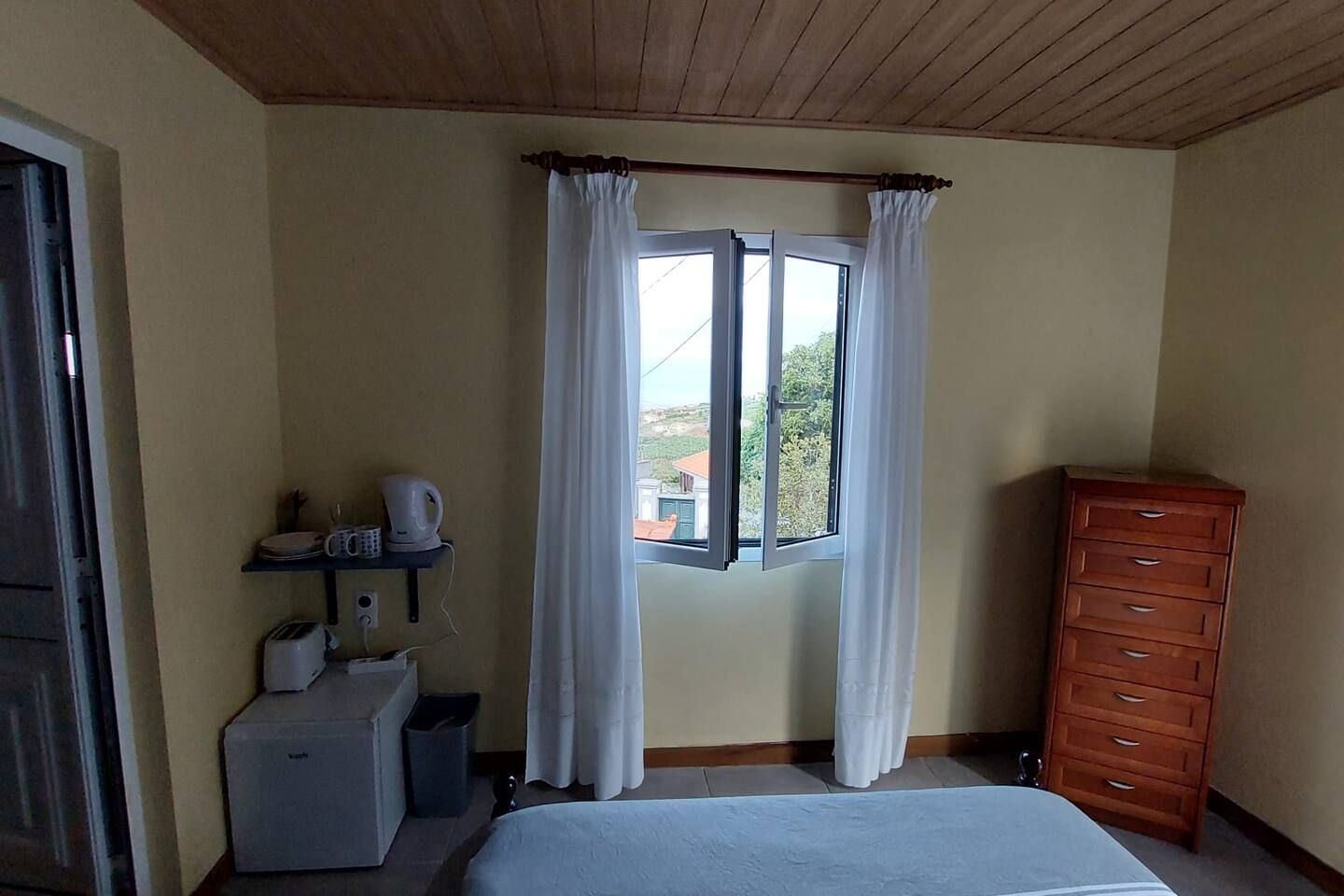 A bedroom with a bed, a dresser, a window with curtains, a shelf with a kettle and a cup, and a small table with a toaster.