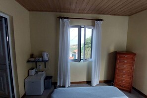 Un dormitori amb un llit, una petita còmoda, una finestra amb cortines, una estant amb una tetera i una tassa, i una taula petita amb una torradora.