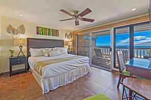A bedroom with a bed, a nightstand, a lamp, a ceiling fan, and a view of the beach through a sliding glass door.