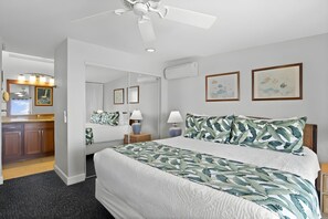 A hotel room with a bed, a nightstand, a lamp, a ceiling fan, a bathroom with a mirror, and artwork on the walls.