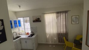 A compact kitchen with a sink, stove, and cabinets, a dining area with a yellow chair, and a large window with curtains.