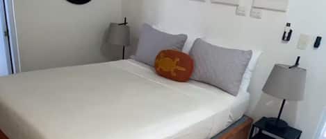 A bedroom with a wooden bed frame, a white mattress, a round wall decoration, a small bedside table with a lamp, and a yellow rug.