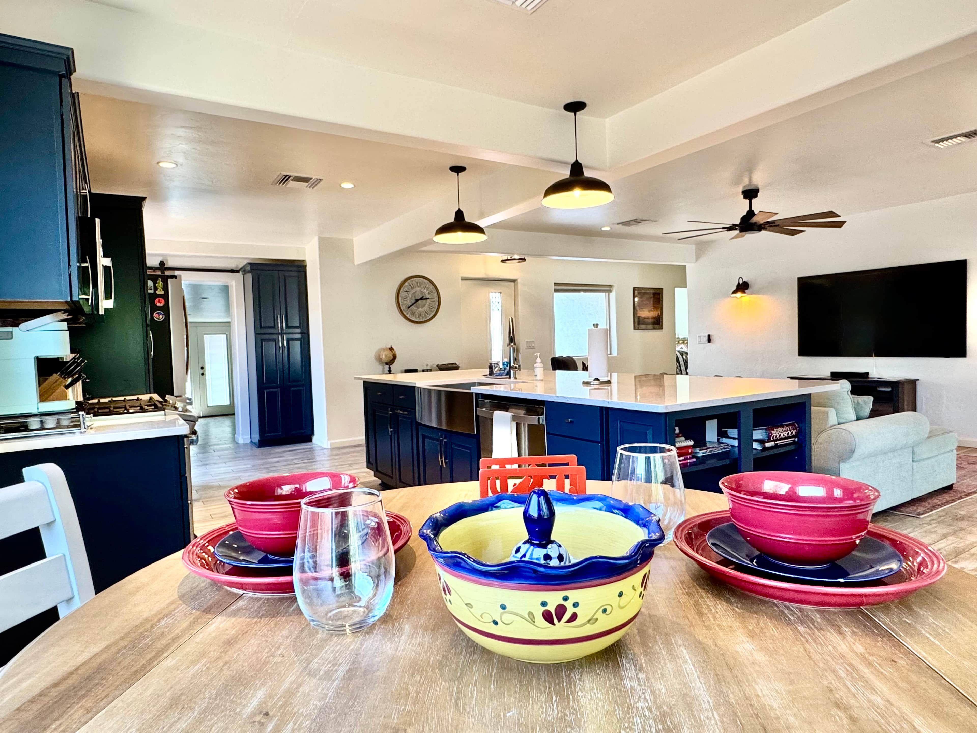 A modern kitchen with a dining area, featuring a central island, pendant lights, and a large flat-screen TV mounted on the wall.