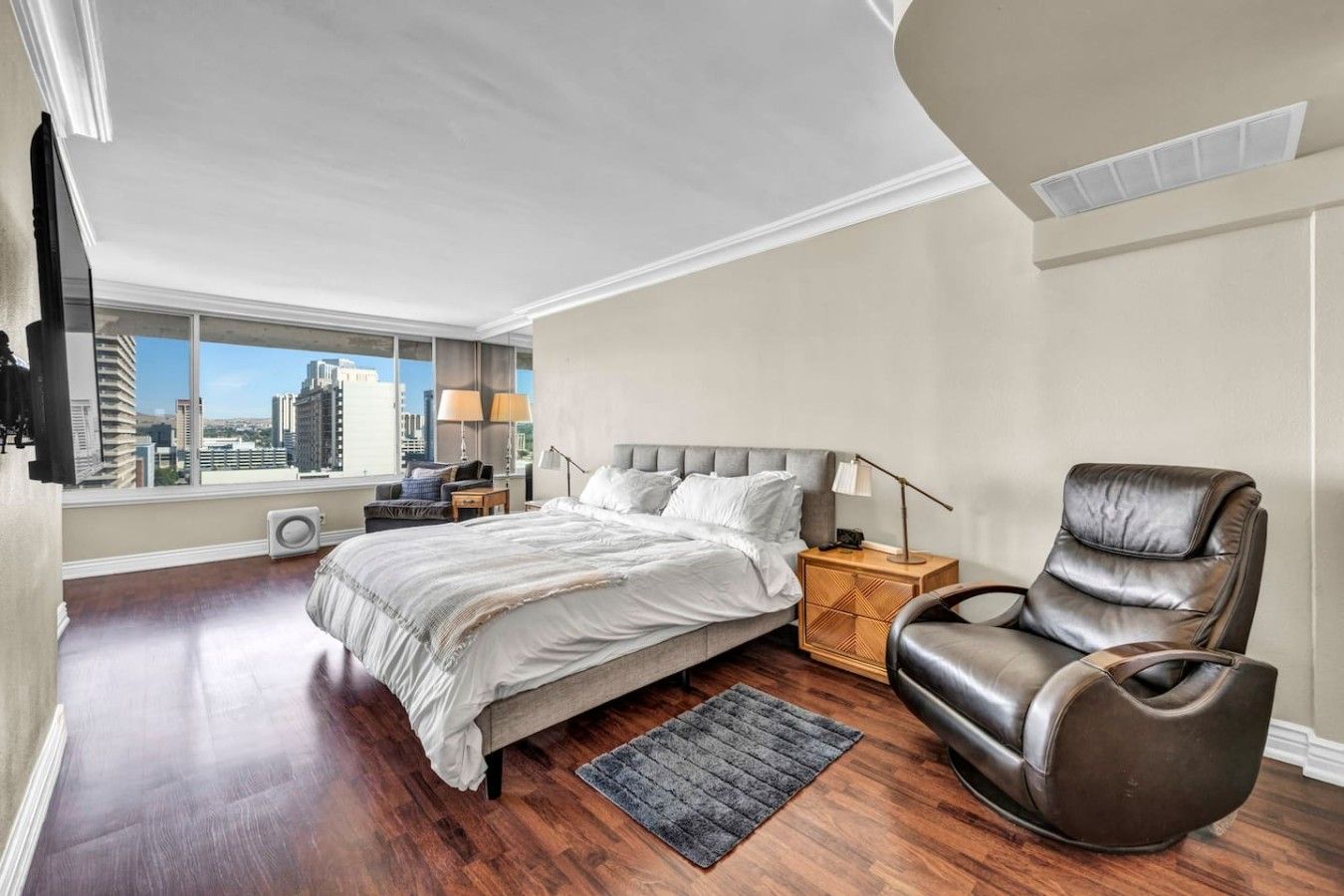 A bedroom with a large bed, a nightstand, a leather chair, and a view of the city through a large window.