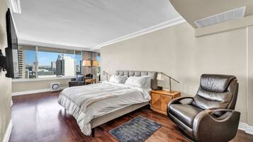 A bedroom with a large bed, a nightstand, a leather chair, and a view of the city through a large window.