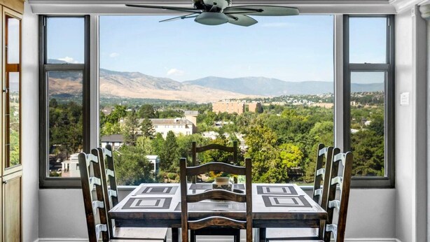 A dining area with a table and chairs, large windows with a view of a town and mountains, and a ceiling fan. - Reno Penthouse | Private Balcony | Mountain Views (Reno)