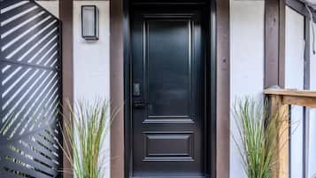 A black door with a keypad lock, flanked by two potted plants, and a metal gate with diagonal slats to the left.