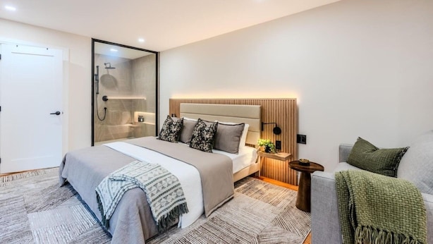 A modern bedroom with a large bed, a bedside table with a lamp, a nightstand, a grey sofa, and a walk-in closet.