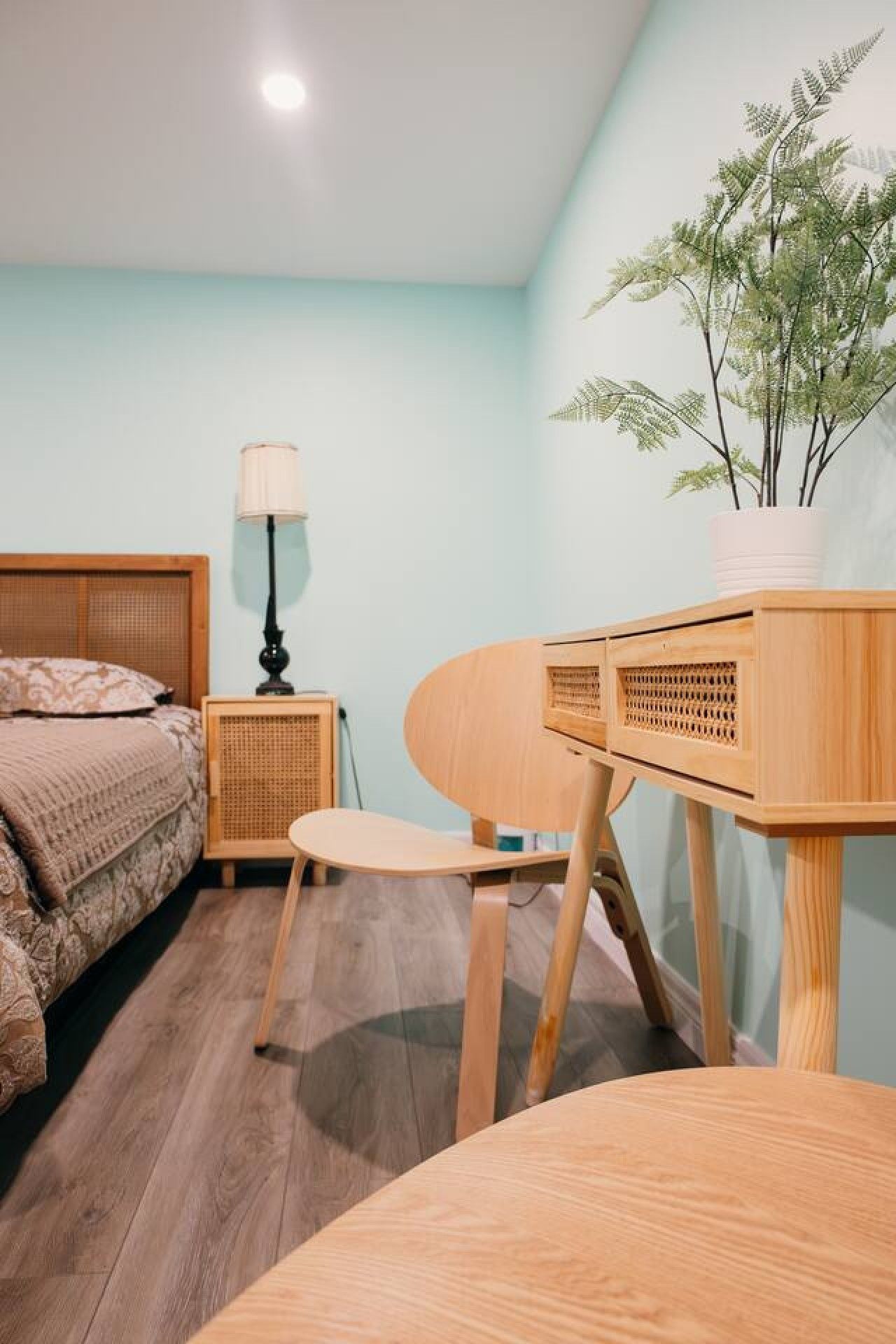 Un dormitorio con cama, mesita de noche, lámpara, silla y un pequeño escritorio con una planta.