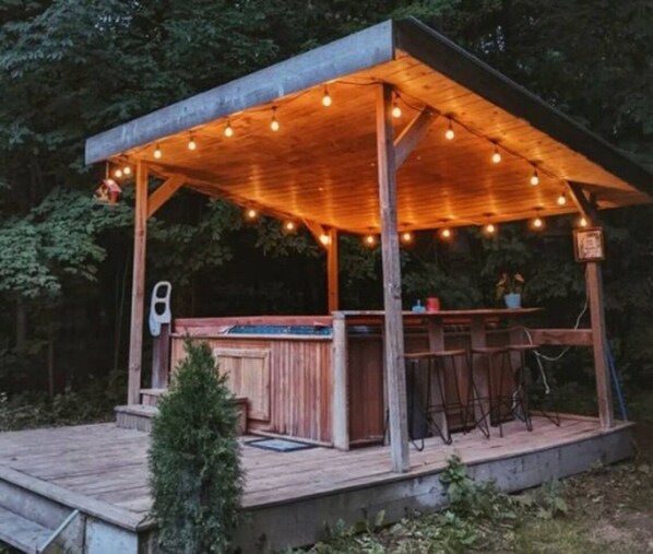 Un espace bar extérieur en bois, doté d’un coin salon abrité et illuminé par des guirlandes lumineuses.