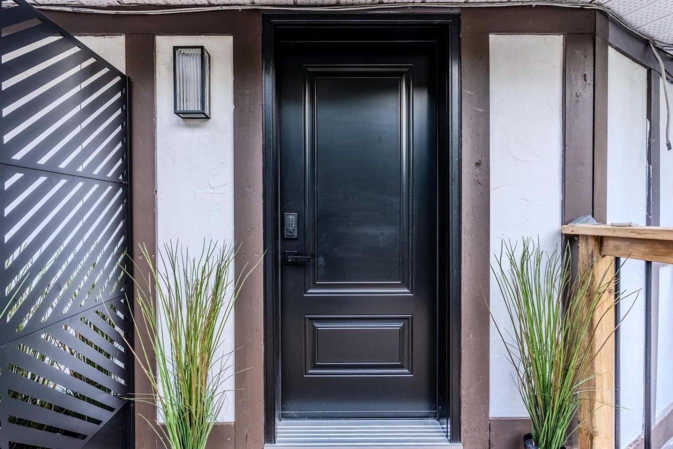 Una puerta negra con cerradura electrónica, flanqueada por dos macetas y una reja metálica con listones diagonales a la izquierda.