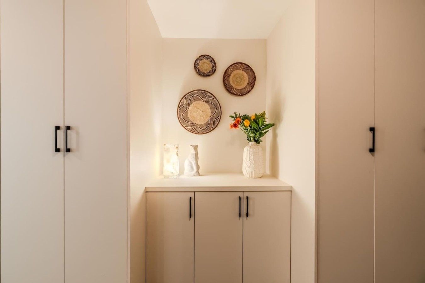 A room with a light-colored wardrobe, a shelf with decorative plates, a vase with flowers, and a vase with a marble-like object.