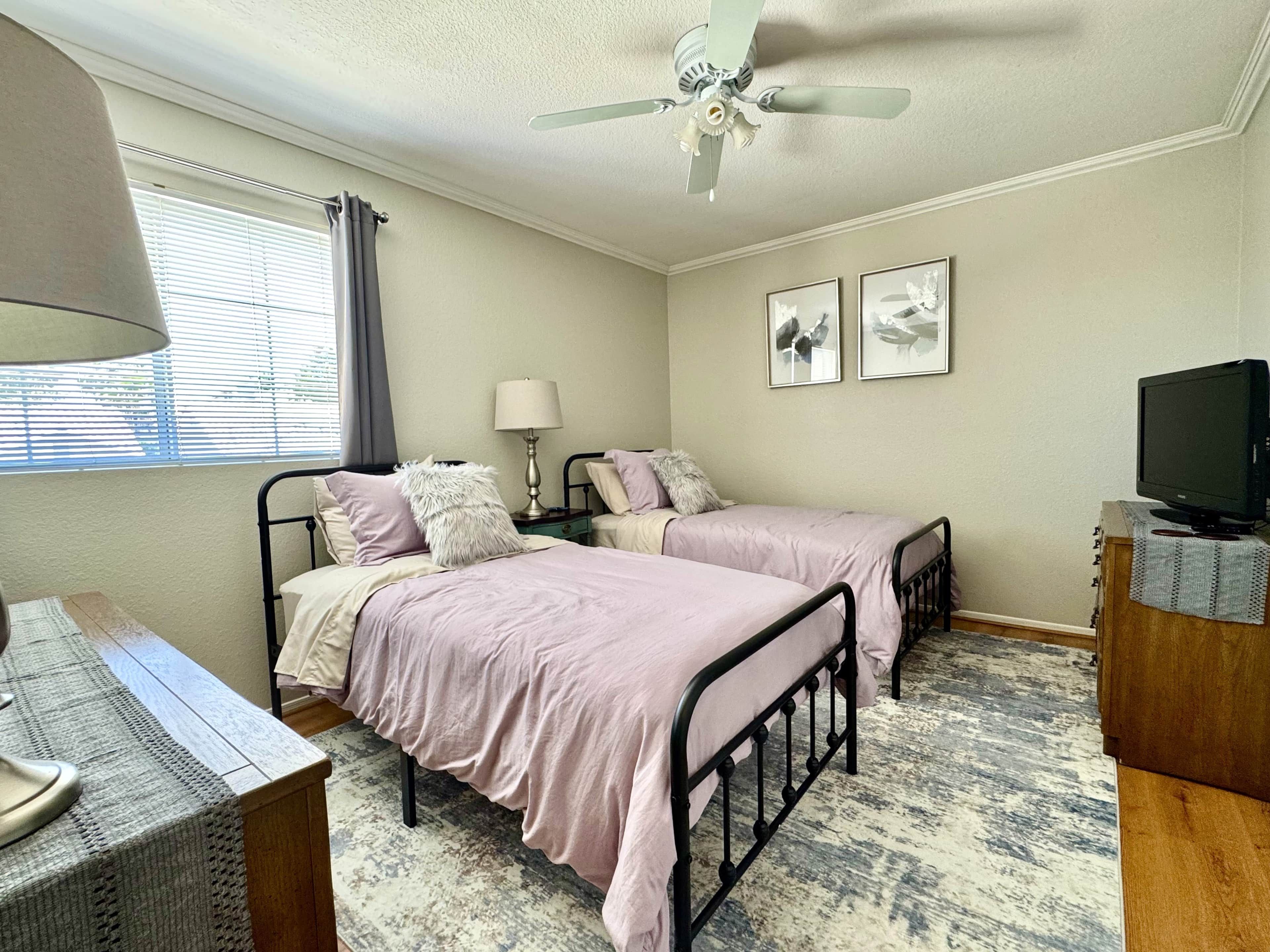 A bedroom with two beds, a ceiling fan, a bedside table with a lamp, a television, and a window with blinds.