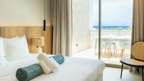 Habitación de hotel con cama, mesita de noche, lámpara, vista al océano a través de un ventanal grande y balcón con mobiliario exterior.