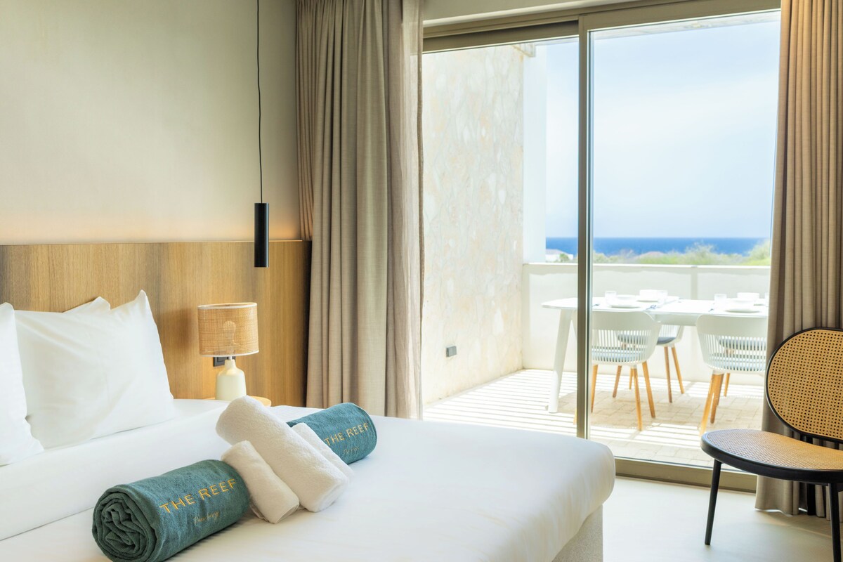 A hotel room with a bed, a nightstand, a lamp, a view of the ocean through a large window, and a balcony with outdoor furniture.