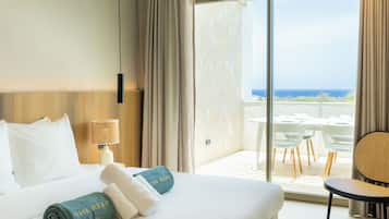 A hotel room with a bed, a nightstand, a lamp, a view of the ocean through a large window, and a balcony with outdoor furniture.