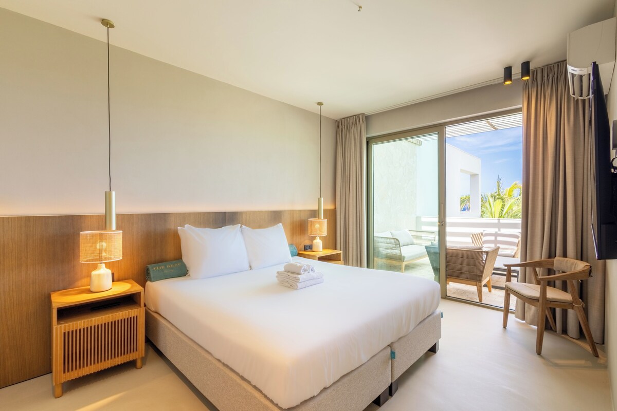 A hotel room with a large bed, a nightstand, a lamp, a window with curtains, and a view of the outdoors.