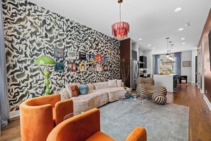 Ein modernes Wohnzimmer mit einer Wand mit Muster, einem Ecksofa, orangefarbenen Sesseln und einem Essbereich im Hintergrund.