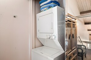 En tvättmaskin med frontmatning, ett kontrollpanel, staplade handdukar och ett träskåp med en stol.