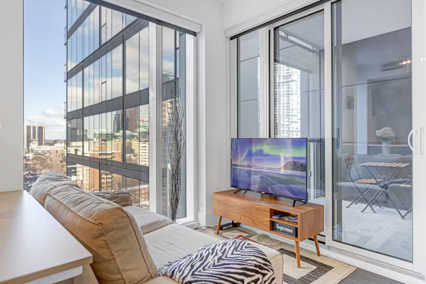 A modern living room with a sofa, a flat-screen TV on a wooden stand, and a city view through large windows.