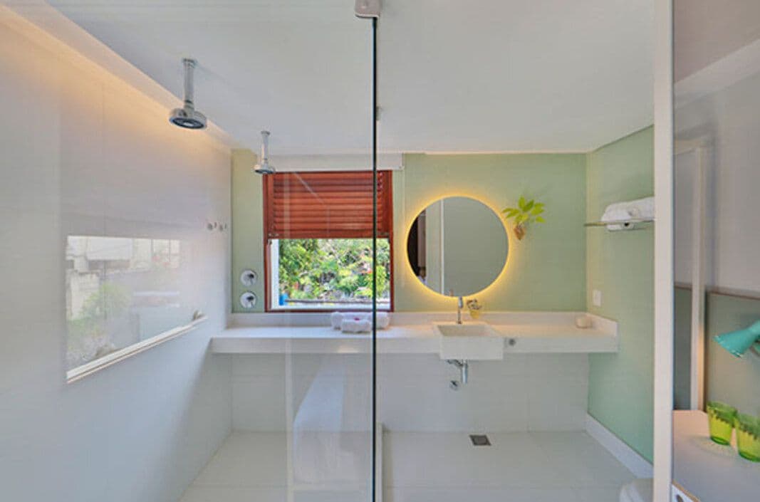 Un baño moderno con cabina de ducha de vidrio, un espejo redondo, un lavabo blanco y una ventana con persianas.