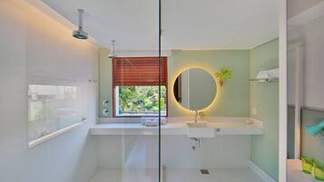 Un baño moderno con cabina de ducha de vidrio, un espejo redondo, un lavabo blanco y una ventana con persianas.