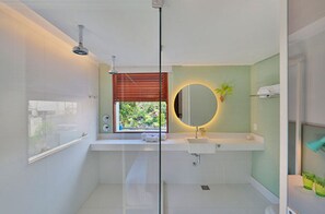 A modern bathroom with a glass shower enclosure, a round mirror, a white sink, and a window with blinds.