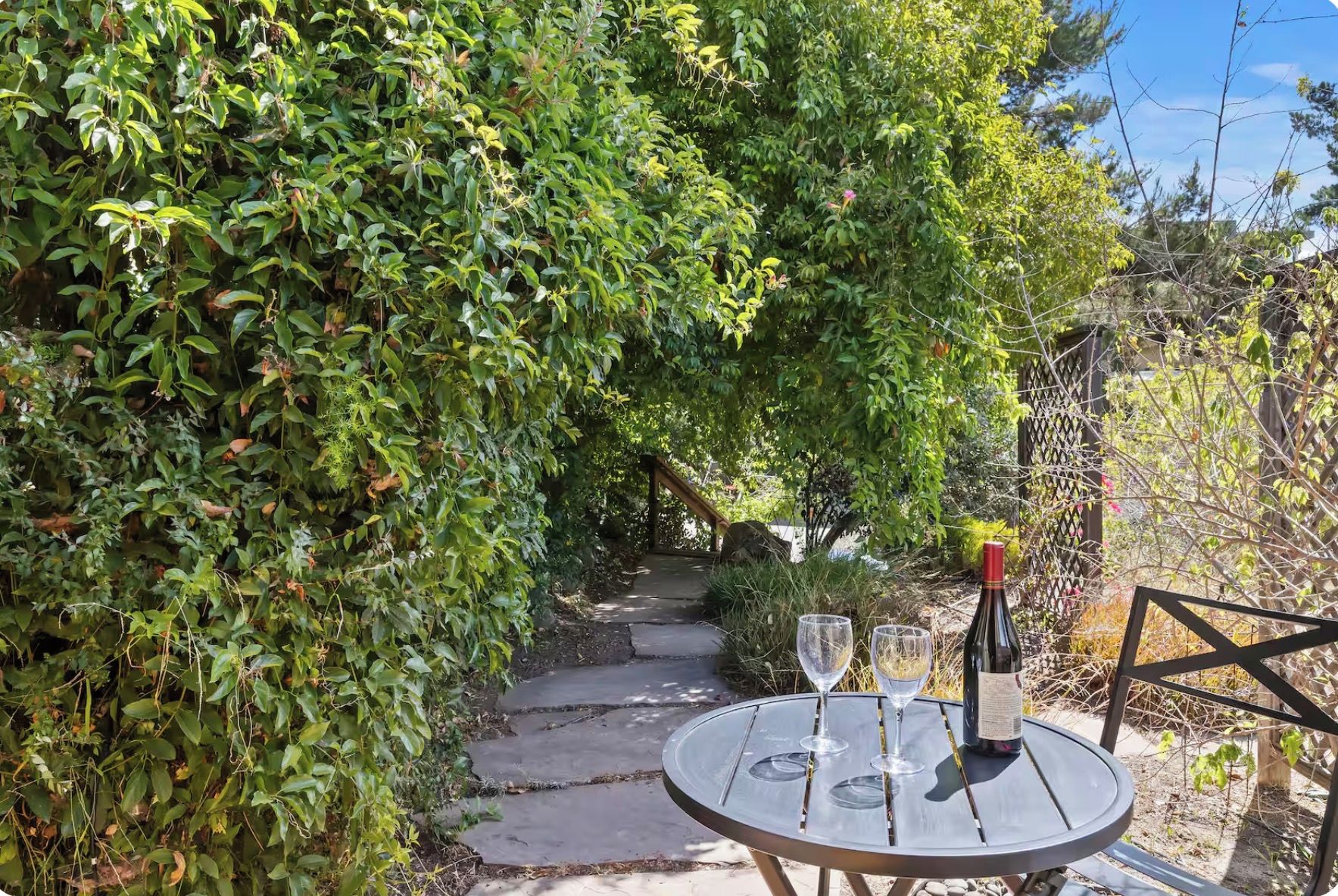 Un chemin de jardin qui mène à une table sur laquelle se trouvent deux verres à vin et une bouteille.