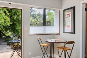 Une pièce avec une fenêtre donnant sur un espace verdoyant, une petite table avec des chaises et une œuvre d’art encadrée accrochée au mur.