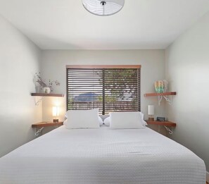 Une chambre à coucher avec un lit, une fenêtre avec des stores, des lampes de chevet et des objets décoratifs sur des étagères.