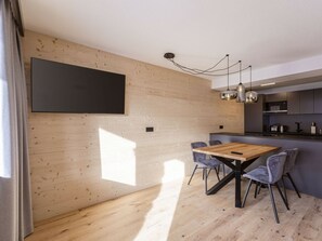 Uma cozinha moderna com armários de madeira, área de refeições com mesa e cadeiras de madeira, e uma grande televisão de tela plana na parede.