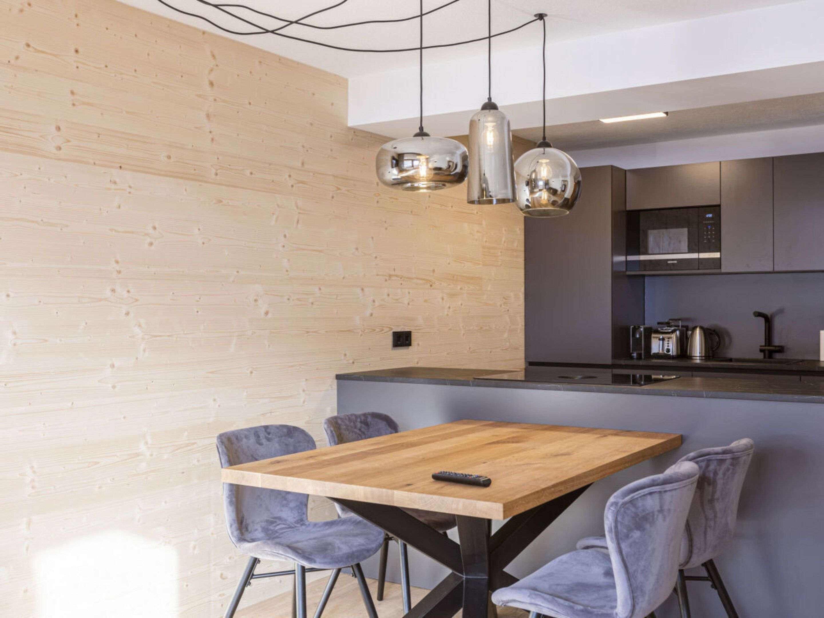 A modern kitchen with a wooden island, pendant lighting, and grey upholstered chairs.