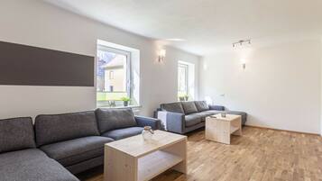A modern living room with a sectional sofa, a coffee table, and a smaller sofa. There are windows providing natural light and views of the outside.