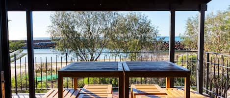 A covered outdoor seating area with wooden tables and benches, overlooking a beach and the sea.