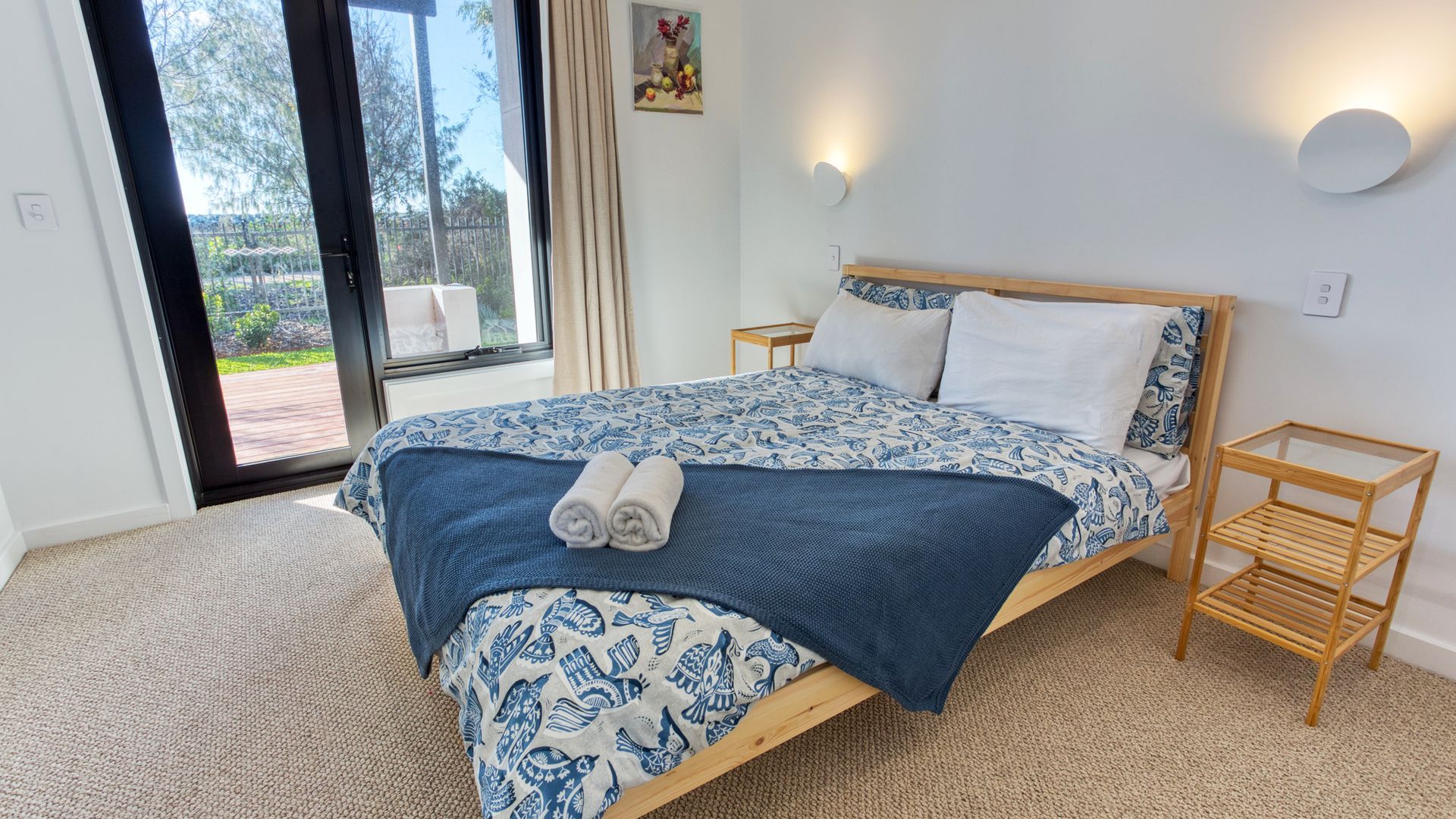 A bedroom with a large bed, a wooden nightstand, a glass coffee table, and a view of the outdoors.