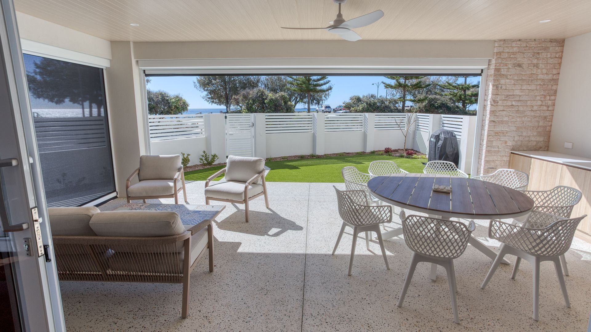 An open patio with a table and chairs, a sofa, and a view of the sea through a glass door.